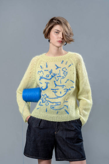 Woman wearing lime coloured mohair sweater and carrying blue wool cone under the arm. The sweater is adorned with sky blue abstract embroidered shapes.